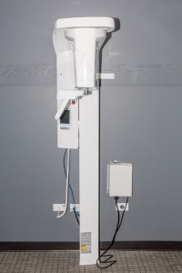 Tall white dental X-ray unit with vertical arm, control box, and power outlets on gray wall