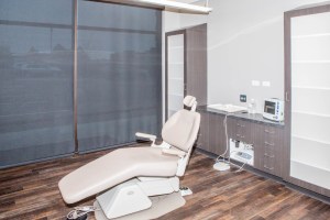 Treatment room with beige dental chair, large window with blinds, cabinets, and sink station