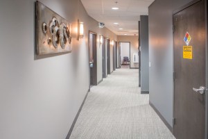 Office hallway with carpeted floor, wall art, closed doors, and fire safety sign on right door