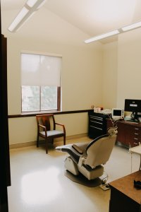 Dental exam room: chair, wooden accent chair, window with blind, desk, and computer monitor